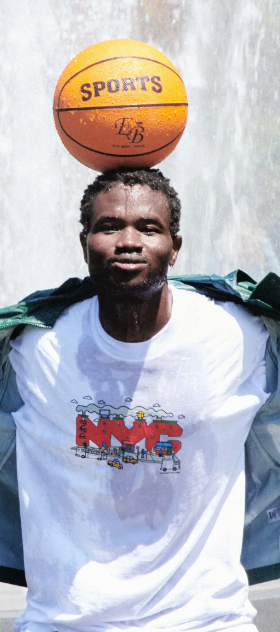 A man standing in a sprinkler with a basketball balanced on his head wearing Only NY t-shirt and jacket