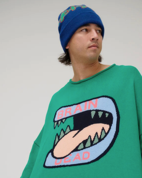 A man wearing a green Brain Dead Sweater with a drawing of a monsters mouth with it's tongue out on it and a blue beanie