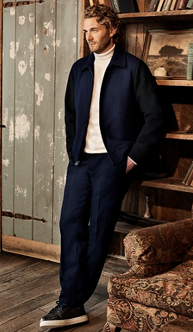 A man with blonde curly hair in a lodge cabin waring a navy blue Club Monaco jacket and pants, with a beige turtleneck