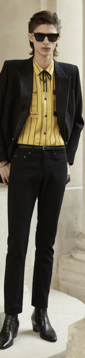 A slim man wearing a Yves St Laurent yellow striped button up with a bolo tie, black blazer, pants and shoes.