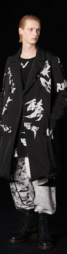 A man with long slicked back hair wearing a Yohji Yamamoto black trenchcoat with white brush strokes across it and black boot