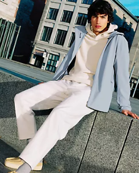 A man sitting wearing a Lululemon outfit feating a light blue jacket, cream hoodie and white pants
