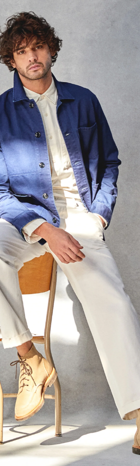 A man sitting on a chair while wearing a Todd Snyder outfit with a denim overcoat and cream pants