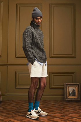 A man in a New Balance outfit with a dark grey fisherman sweater, a matching beanie and blue socks
