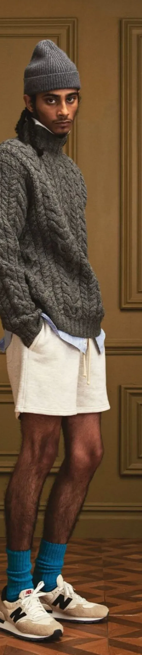 A man in a New Balance outfit with a dark grey fisherman sweater, a matching beanie and blue socks