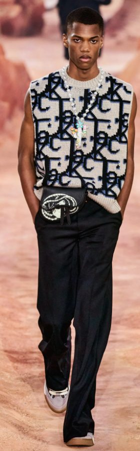 A man wearing a Dior allover print vest, black pants and a colorful cross necklace