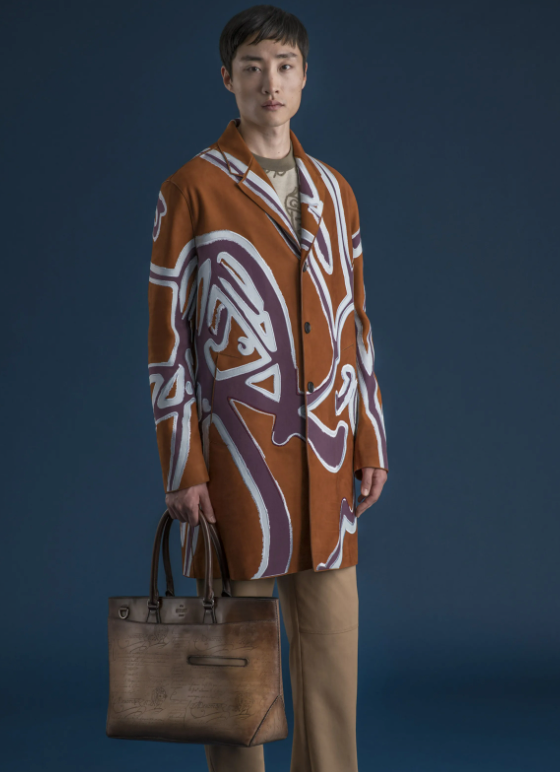 A man wearing a long Berluti jacket with a large purple pattern on top and brown pants