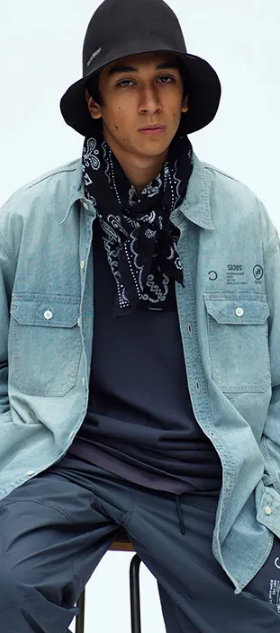 A man sitting in a Neighborhood outfit feating a navy bandana, denim overshirt, technical pants and a bucket hat