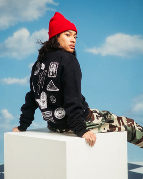 A woman sitting and wearing a 10 Deep hoodie with camo pants and a red beanie