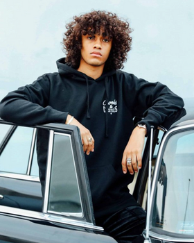A man wearing a Crooks & Castles hoodie standing beside a black car