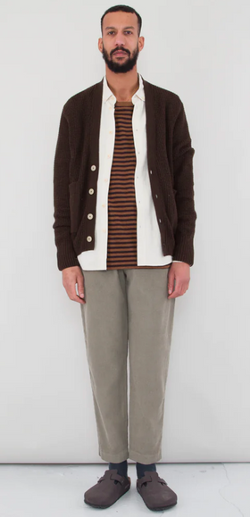 A man wearing a Folk Clothing layered outfit with a brown cardigan and white button up and grey pants