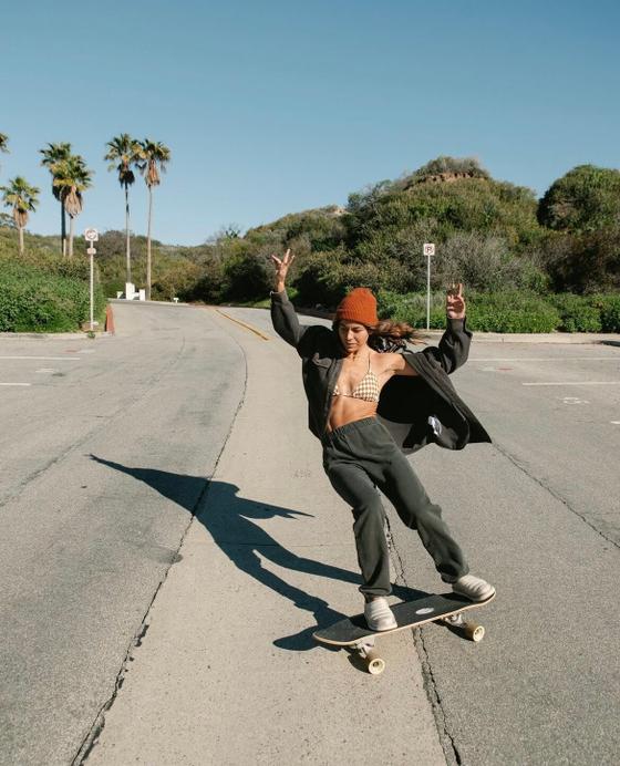 A woman wearing Sanuk slip-ons while skateboarding down a palm-lined coastal road