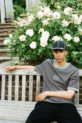 A man wearing a grey Yardsale ringer T-shirt and black cap sitting on a park bench